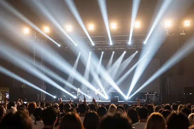 View from a concert crowd looking toward a lit stage with sweeping white light beams