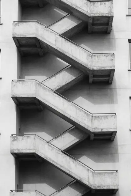 Black and white close-up of a zigzagging external concrete staircase on an apartment building in sharp noon light