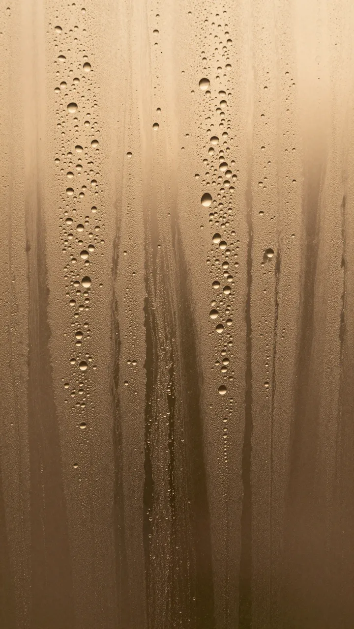 Close-up of water droplets and streaky condensation on glass lit by warm amber light behind