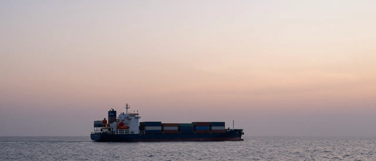 A loaded container ship sails calm ocean water beneath a soft hazy pastel sunrise sky