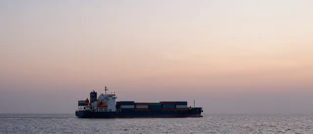 A loaded container ship sails calm ocean water beneath a soft hazy pastel sunrise sky