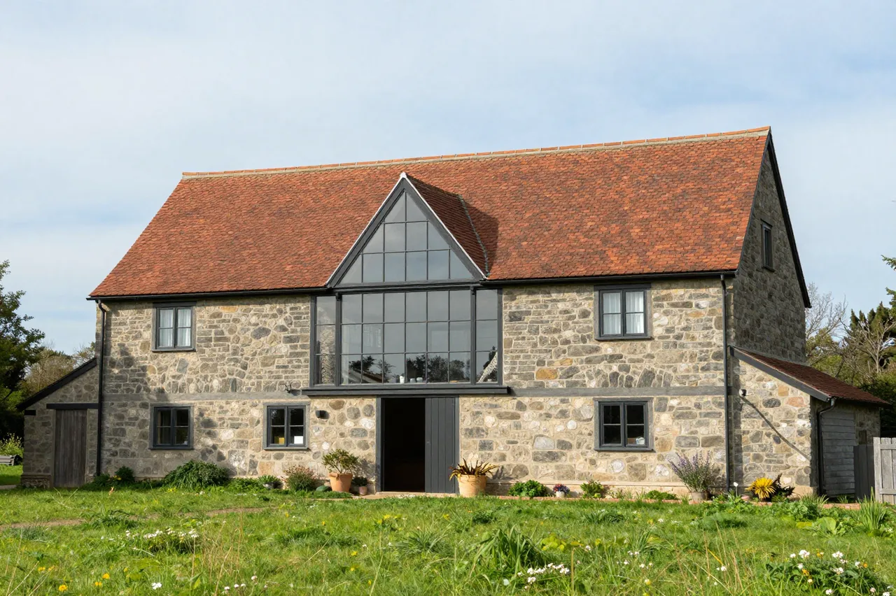 A stone converted barn home with a glass gable sits in a grassy wildflower meadow under a pale sky