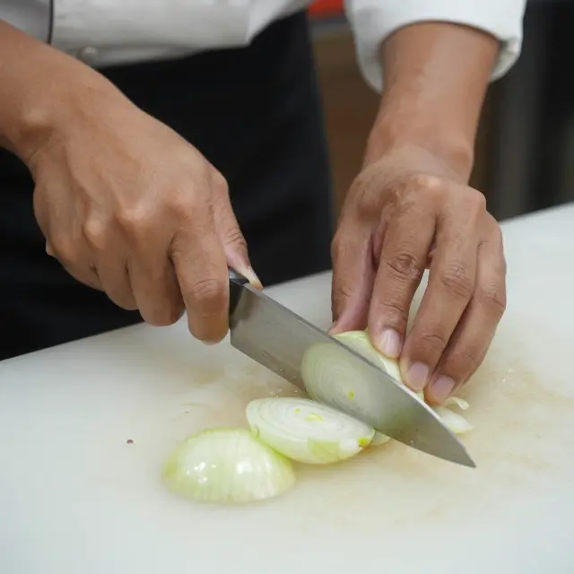 Close-up of a line cook's hands slicing a white onion with a chef's knife on a pale cutting board.