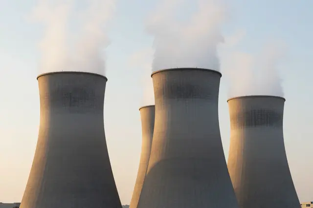 Four concrete power plant cooling towers release steam into a pale warm sunrise sky