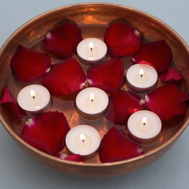 Top view of a copper bowl holding floating red rose petals and lit tealight candles on a smooth gray surface