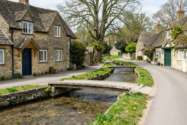 A quiet village lane runs alongside a clear millstream crossed by small stone footbridges between stone cottages in spring.