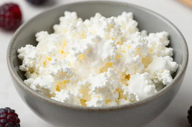 A close-up of a gray ceramic bowl filled with cottage cheese, surrounded by fresh blackberries on a light table surface.