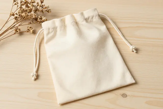 Blank natural cotton drawstring pouch rests on light wood beside delicate dried flowers.