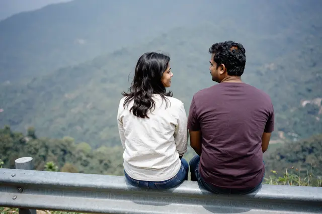 A South Asian couple sits on a metal guardrail at a mountain overlook, facing each other with a forested mountain range