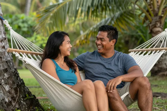 A Pacific Islander man and Southeast Asian woman laugh together while sitting in a hammock between palm trees in afternoon