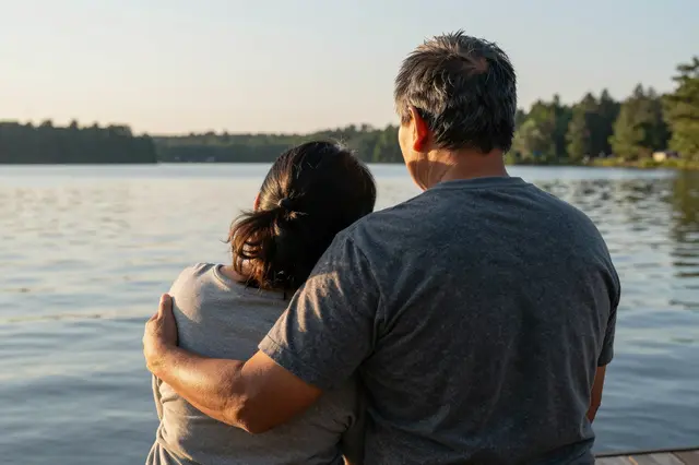 A couple viewed from behind stand embracing on a lakeside dock as the woman rests her head on the man's shoulder at golden