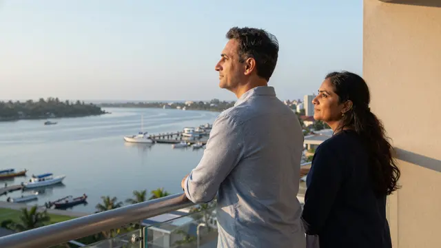 A middle aged man and woman stand on a hotel balcony overlooking a calm harbor at sunrise
