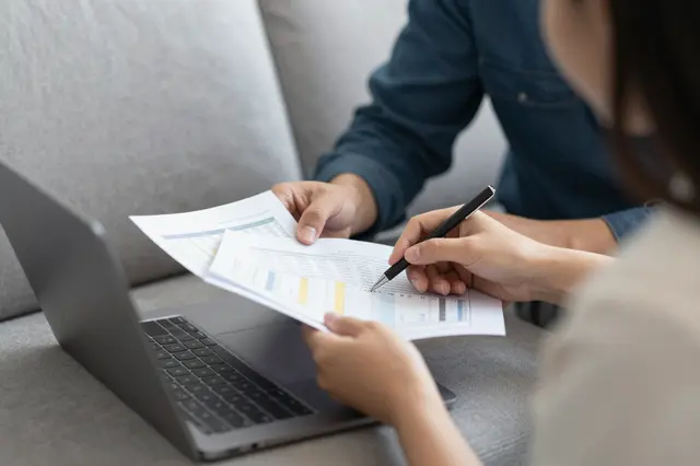 Two people on a gray sofa examine financial documents beside an open silver laptop, one holding a pen.