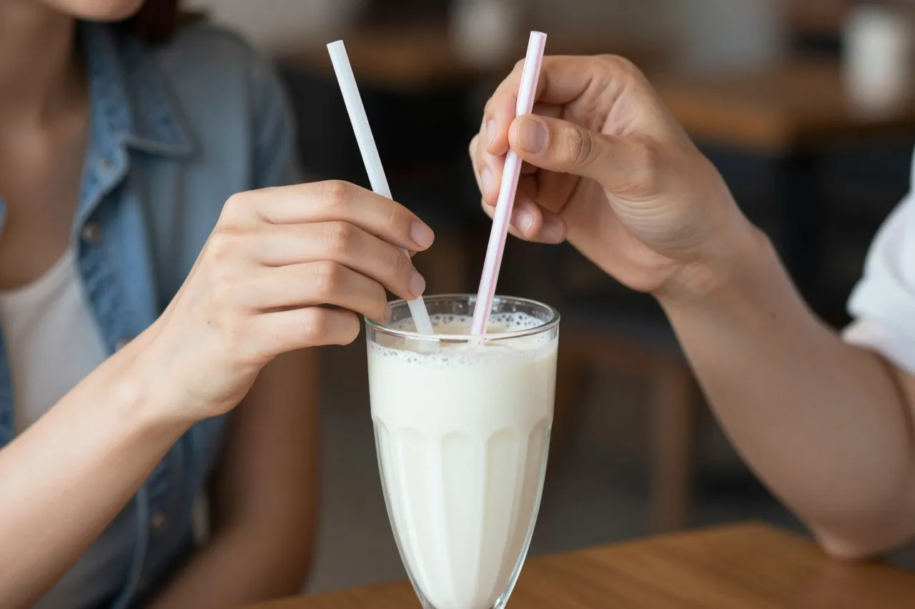 Two people hold straws in a single glass of creamy milkshake on a cafe table.