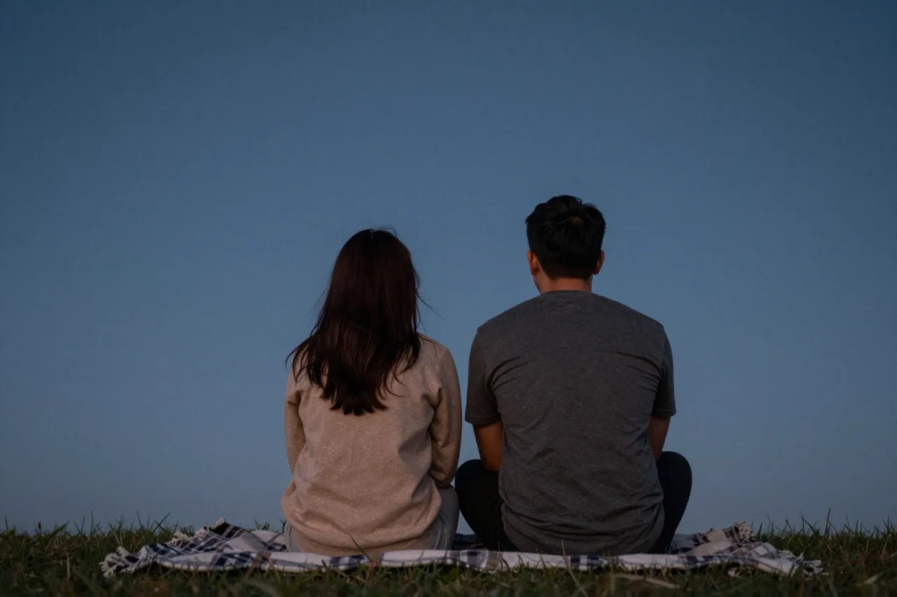 Back view of a couple sitting on a plaid blanket on grass looking out at a pale blue evening sky