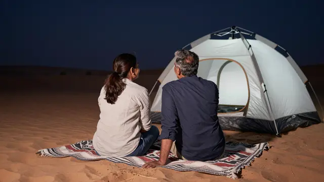 A South Asian couple in their fifties sits from behind on a patterned blanket next to a camping tent in a sandy desert at