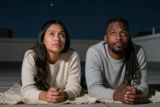 Two adults lie on a fringed rooftop blanket looking up at the dark night sky in plain knit sweaters.