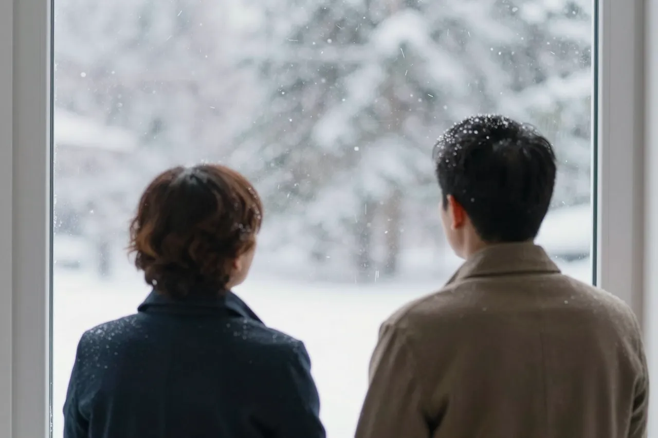 Two adults seen from behind stand indoors looking out a window at falling winter snow