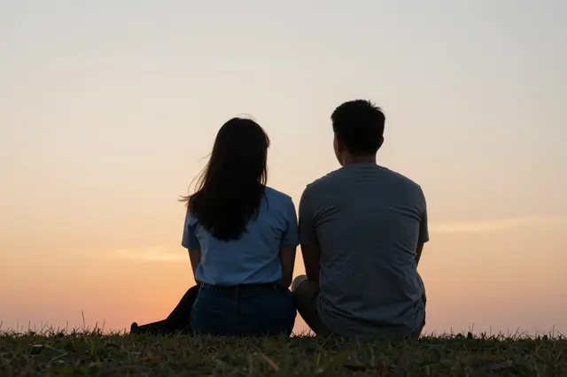 Rear view of a couple sitting on grass atop a hill watching a soft warm sunset sky