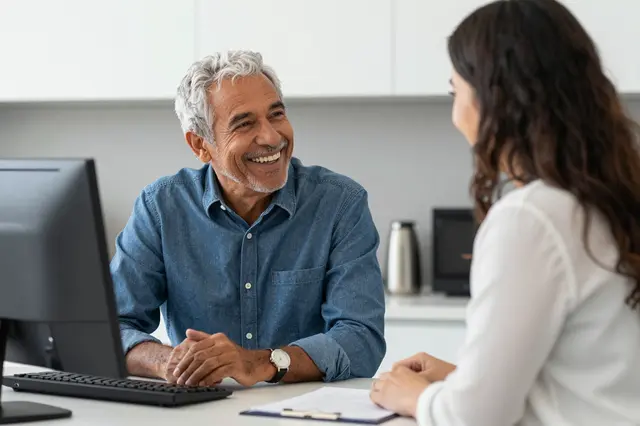 Smiling older mixed-race man converses with a young female colleague at an office desk