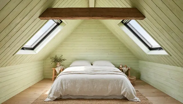 Sunlit attic bedroom with limewashed wood walls, centered bed under twin skylights and a heavy ceiling beam