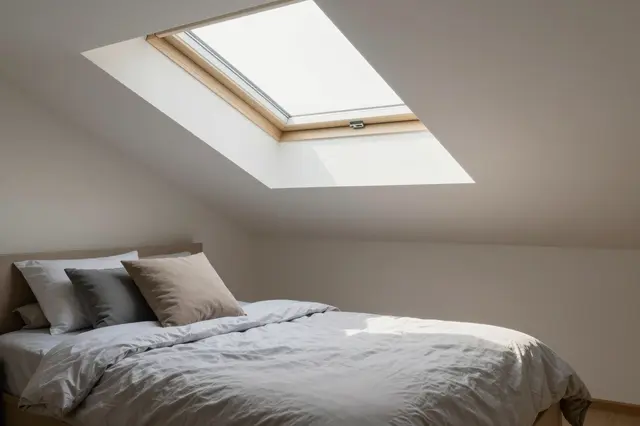 A minimal attic bedroom with a made bed and a wooden-framed skylight in the slanted ceiling