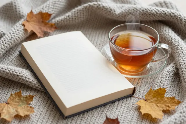 Open book, steaming tea cup, and fall maple leaves rest on a soft knitted blanket