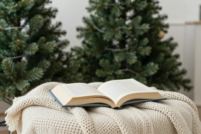 An open hardcover book rests on a cream knitted blanket in front of decorated Christmas trees.