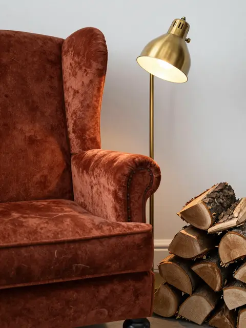Rust velvet wingback chair sits next to a lit brass floor lamp and stack of firewood in a calm interior corner