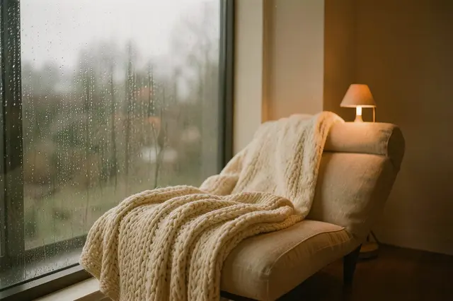 A plush lounge chair with a knit blanket sits beside a rain-streaked window next to a warm glowing table lamp