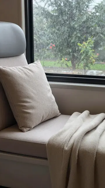 Neutral upholstered window seat with a cushion and knit throw beside a rain streaked window looking out to green foliage