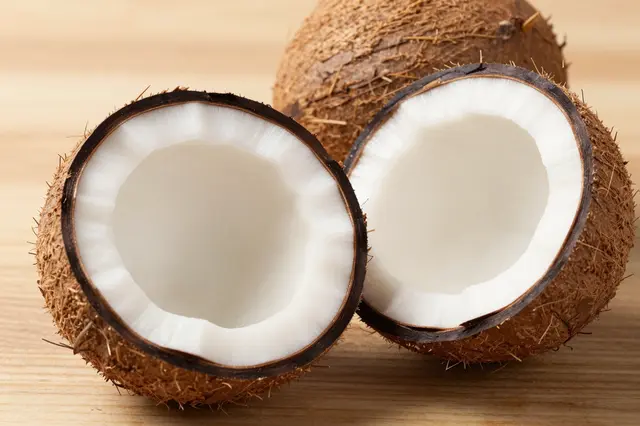 Split coconut halves with white inner flesh rest on a light wooden table beside a whole coconut