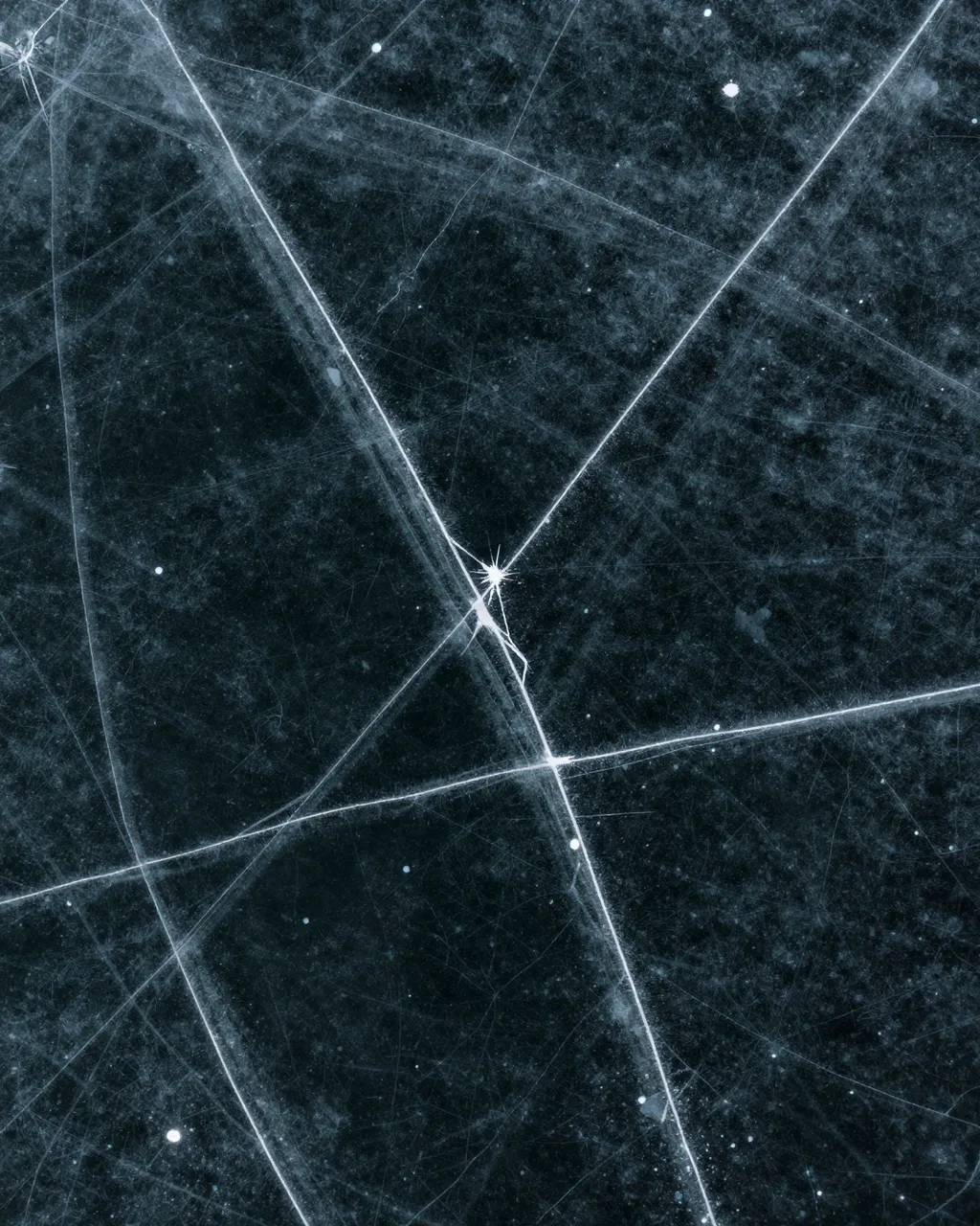 Top-down view of dark black lake ice crisscrossed with thin bright white fracture lines