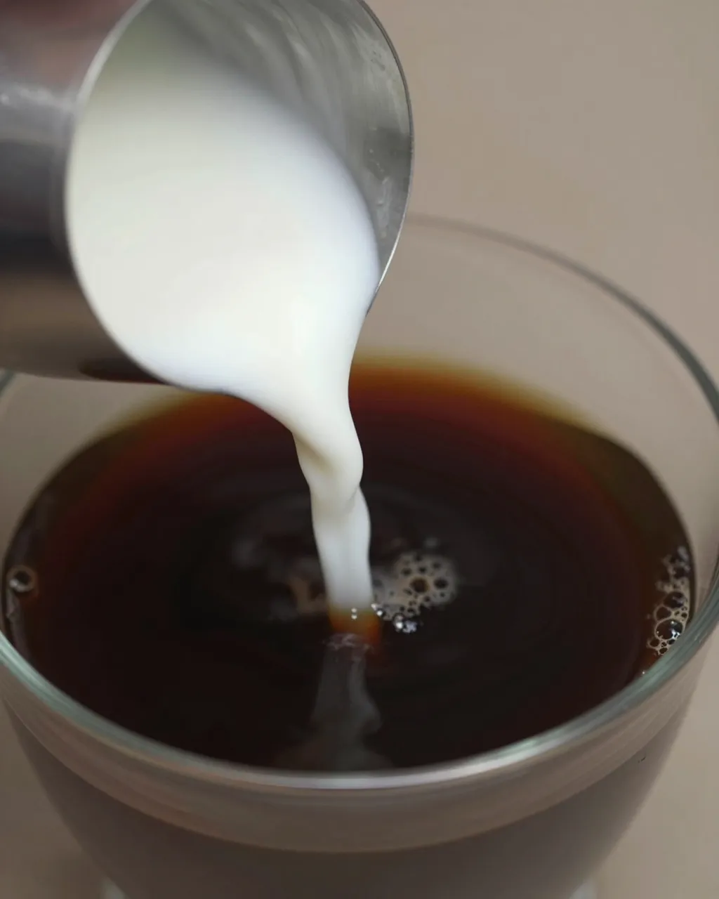 Cream pours from a small metal pitcher into a glass of dark black coffee creating a swirling bubble pattern.