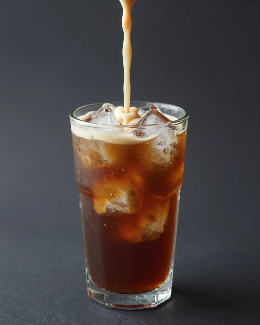 Cream pours into a tall glass of iced cold brew coffee against a dark gray background