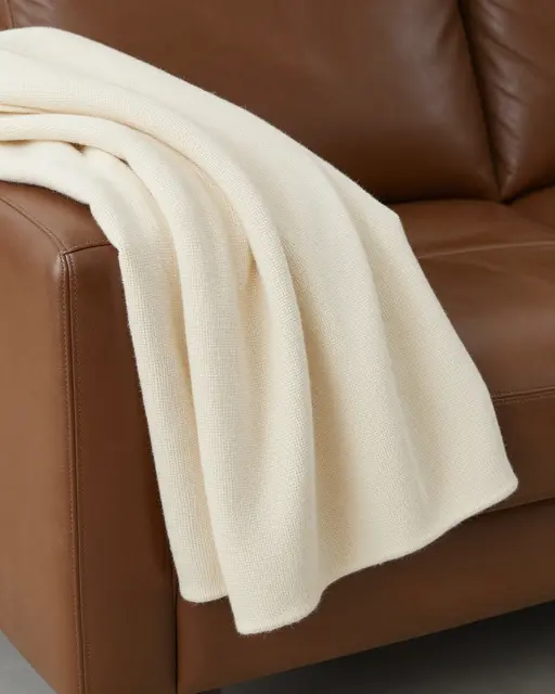 A plain cream woven knit throw blanket draped over the arm of a brown leather sofa