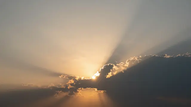 Bright sun peeking behind clouds casts fanning crepuscular rays across a warm sunset sky