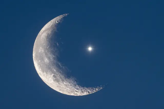 A textured crescent moon sits beside a sharp bright star against a deep blue twilight sky