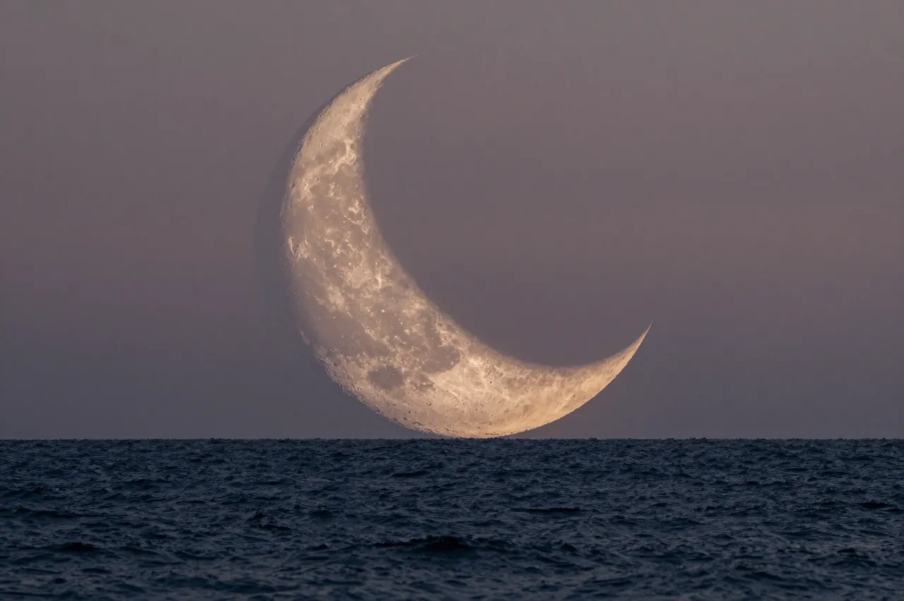 A large detailed crescent moon hangs low above a dark rippled ocean under a pale twilight sky