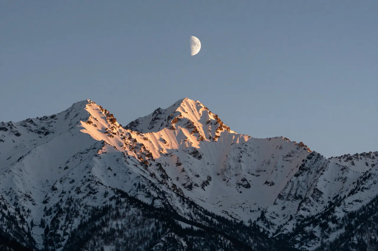 Sunlit snow-covered mountain peaks rise beneath a crescent moon at pale dusk