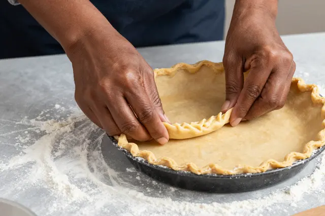 Hands of a Black woman crimping the edge of a raw pie crust on a floured gray table