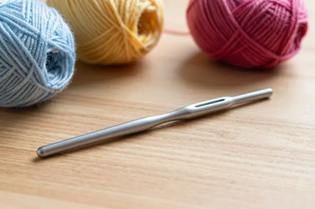 A metal crochet hook rests in front of three colorful yarn balls on a light wooden table.