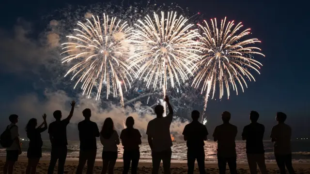 Silhouetted people stand on a beach watching bright golden fireworks burst over the ocean at night