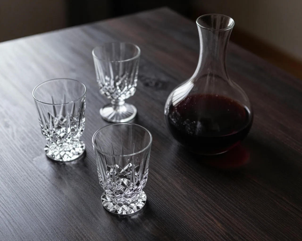 Cut crystal glasses and a red wine decanter rest on a dark wood table in low moody light