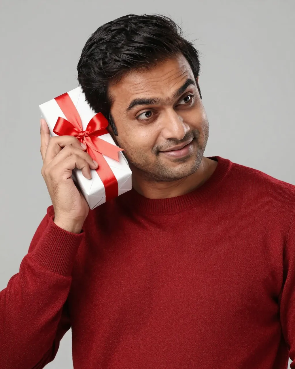 South Asian man in a red sweater holds a wrapped gift beside his ear with a curious smile