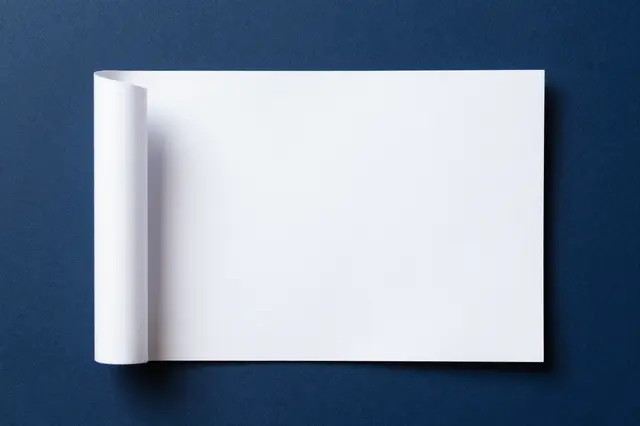 A blank white sheet of paper with a curled left corner rests on a solid dark blue matte background