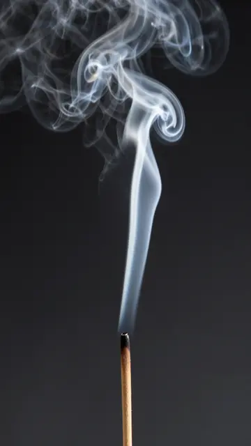 Delicate wispy incense smoke curls upward from a burning stick against a dark black background