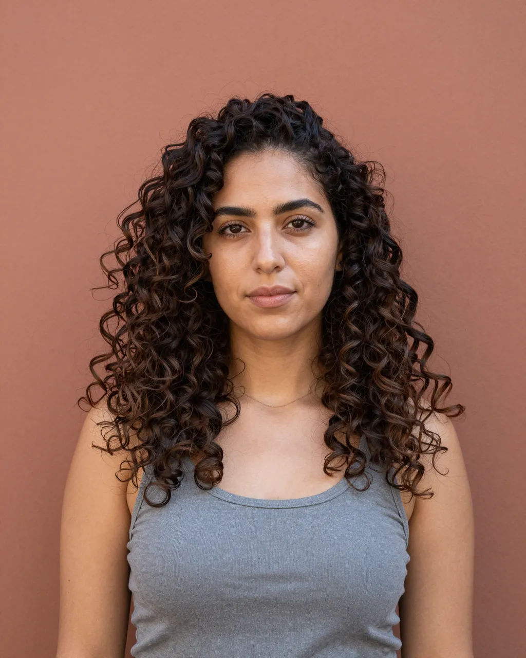 Portrait of a woman in her twenties with curly dark hair standing against a plain terracotta wall
