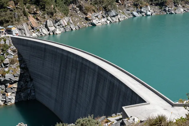 Aerial view of a curved concrete dam holding a turquoise mountain reservoir surrounded by rocky slopes