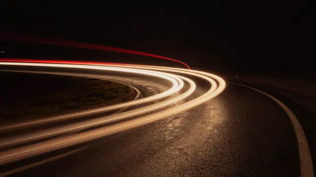 Long exposure red and warm gold light trails curve along a paved winding road after dark